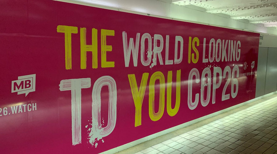 A large pink banner in a public space displaying the text "THE WORLD IS LOOKING TO YOU COP26" in white and yellow letters.