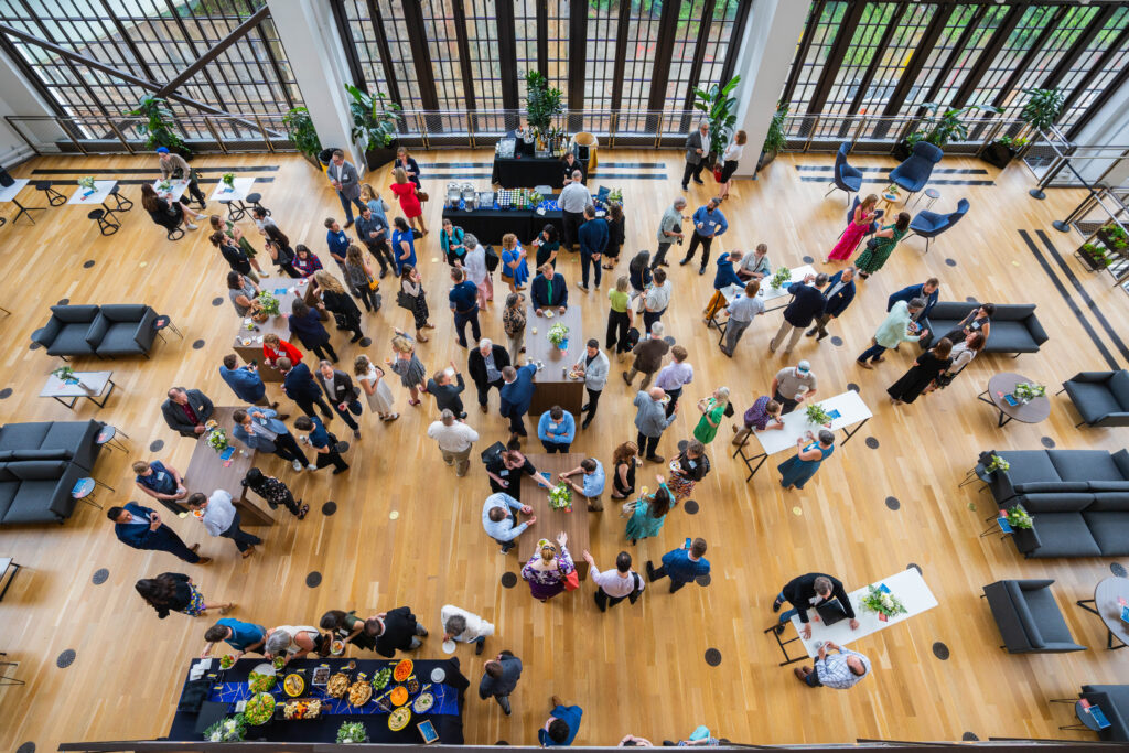2030 Progress Report 2024 attendees from an overhead view of a large group of people socializing in a spacious indoor area with wooden floors and plants. Tables with food and drinks are set up.