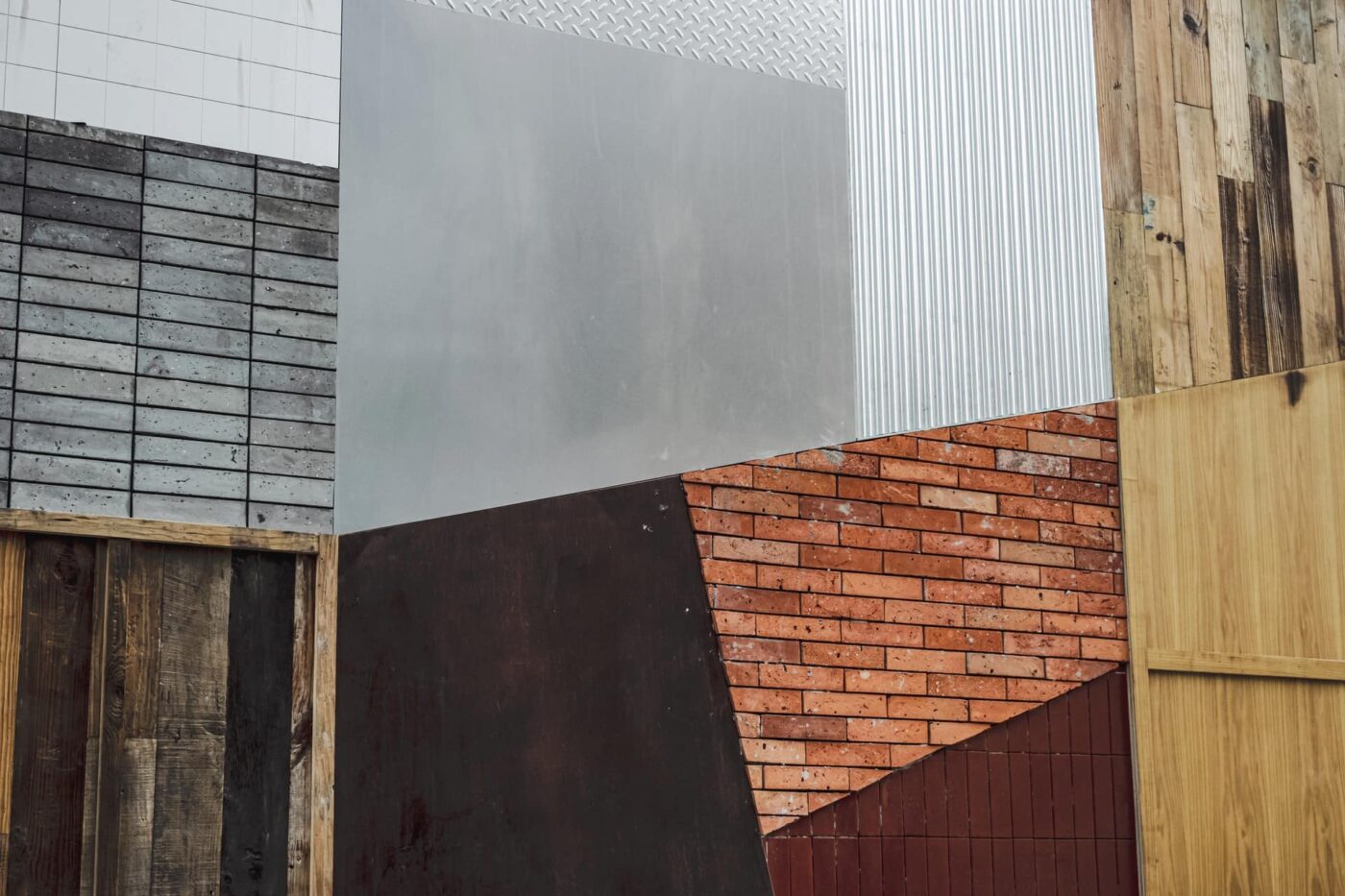 An abstract wall composed of various materials including wood, metal, and brick. The sections are arranged in geometric patterns with different textures and colors, such as gray, brown, and red.