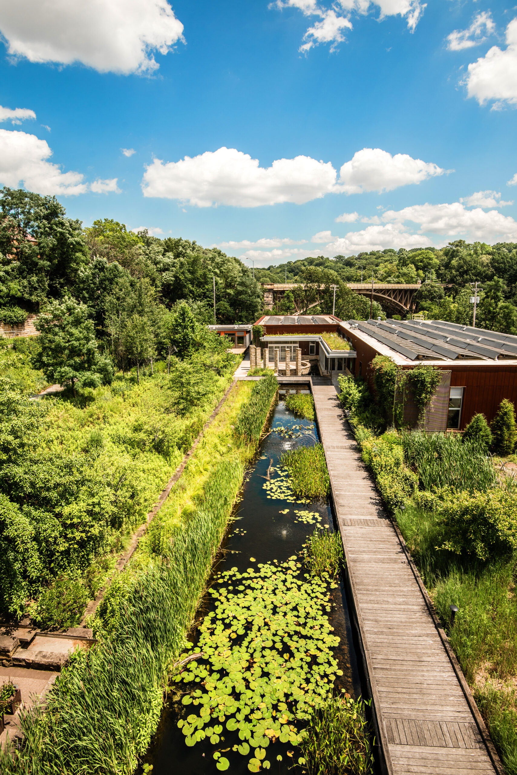 An aerial view of a landscaped area featuring a narrow waterway with lily pads, surrounded by lush greenery, and a wooden walkway leading to a modern building under a bright blue sky with fluffy clouds.