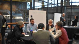 2 women and 3 men having a conversation around a circular table with a whiteboards in the background.