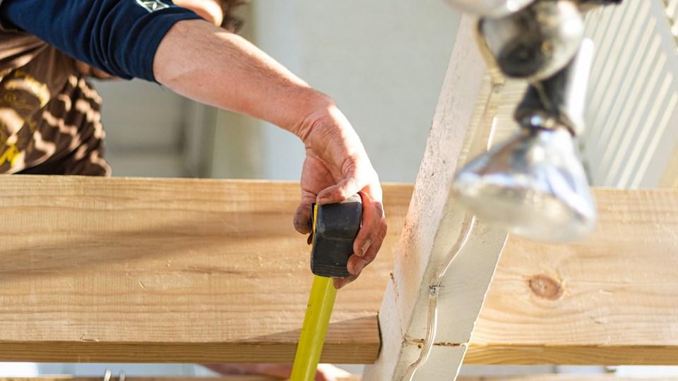 A person's hand measuring a wooden beam with a tape measure. The background includes bright lighting and construction materials.