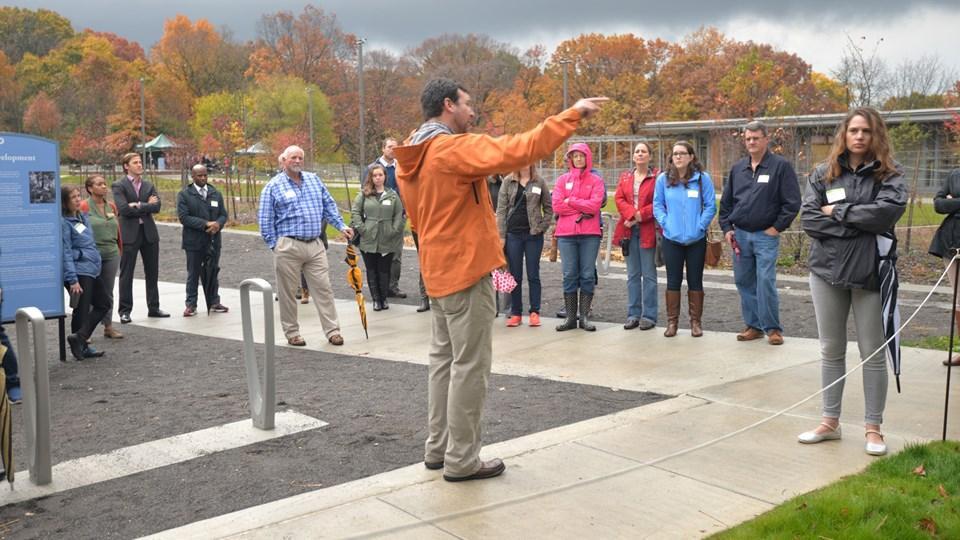 A group of people gathered outdoors, listening to a speaker in an orange jacket. The background features autumn foliage and a cloudy sky. Several attendees are wearing jackets, with some holding umbrellas.